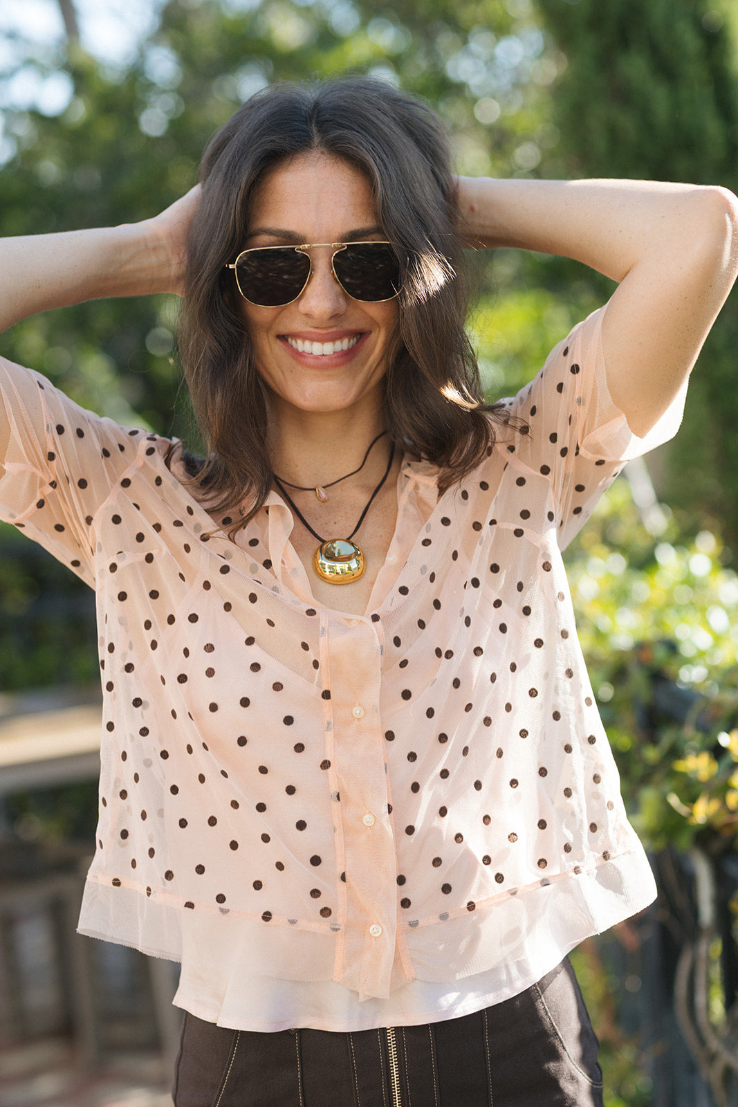 A woman wearing a sheer peach short-sleeve blouse with black polka dots, styled with sunglasses and layered necklaces, posing outdoors with a blurred green background.