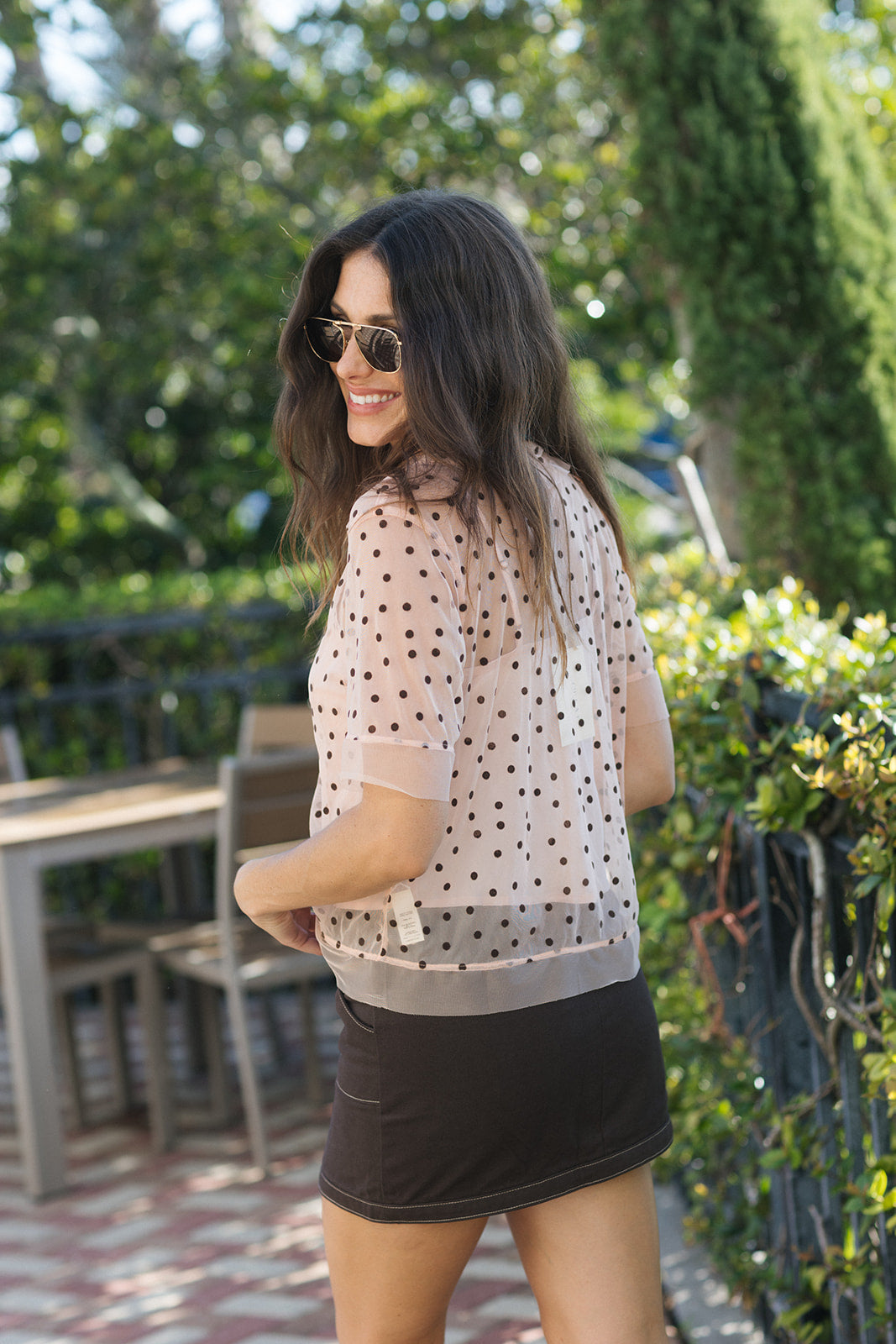A woman wearing a sheer light pink short-sleeve blouse with black polka dots and a black mini skirt, standing outdoors in a garden patio setting.