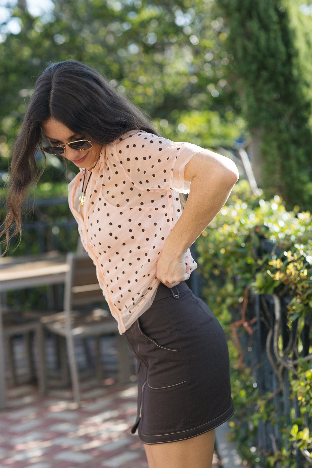 A woman wearing a light pink short-sleeve blouse with black polka dots and a dark brown mini skirt, standing outdoors in a garden setting.