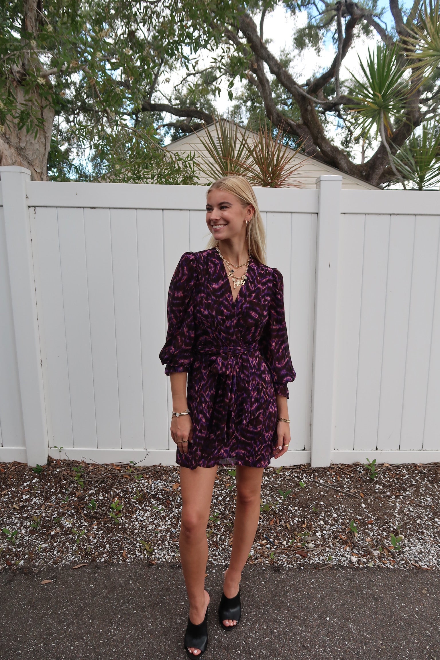 Woman wearing a purple floral dress standing in front of a white fence with trees in the background, full body