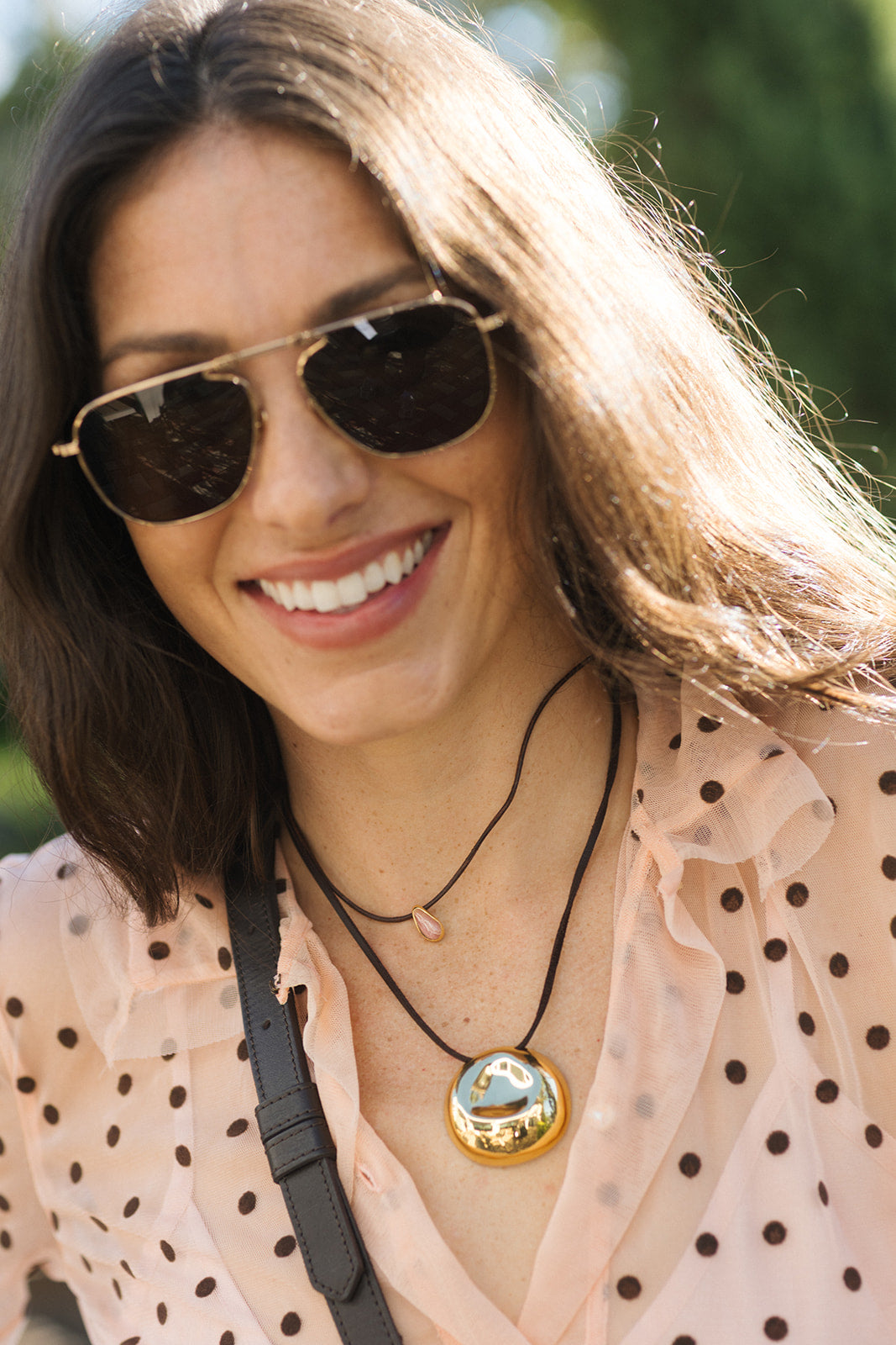 A close-up lifestyle shot of a woman wearing a gold circular pendant necklace with a reflective mirror finish, layered over a black cord necklace and a light pink polka-dot blouse.