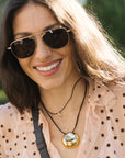 A close-up lifestyle shot of a woman wearing a gold circular pendant necklace with a reflective mirror finish, layered over a black cord necklace and a light pink polka-dot blouse.