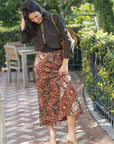 A woman posing outdoors on a brick patio wearing a brown cropped jacket, a rust-orange and black patterned midi skirt, and clear high-heeled sandals, with a brown shoulder bag.