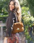 A woman outdoors wearing a brown suede shoulder bag with a gold-tone clasp, styled with a dark jacket and a red floral patterned skirt.