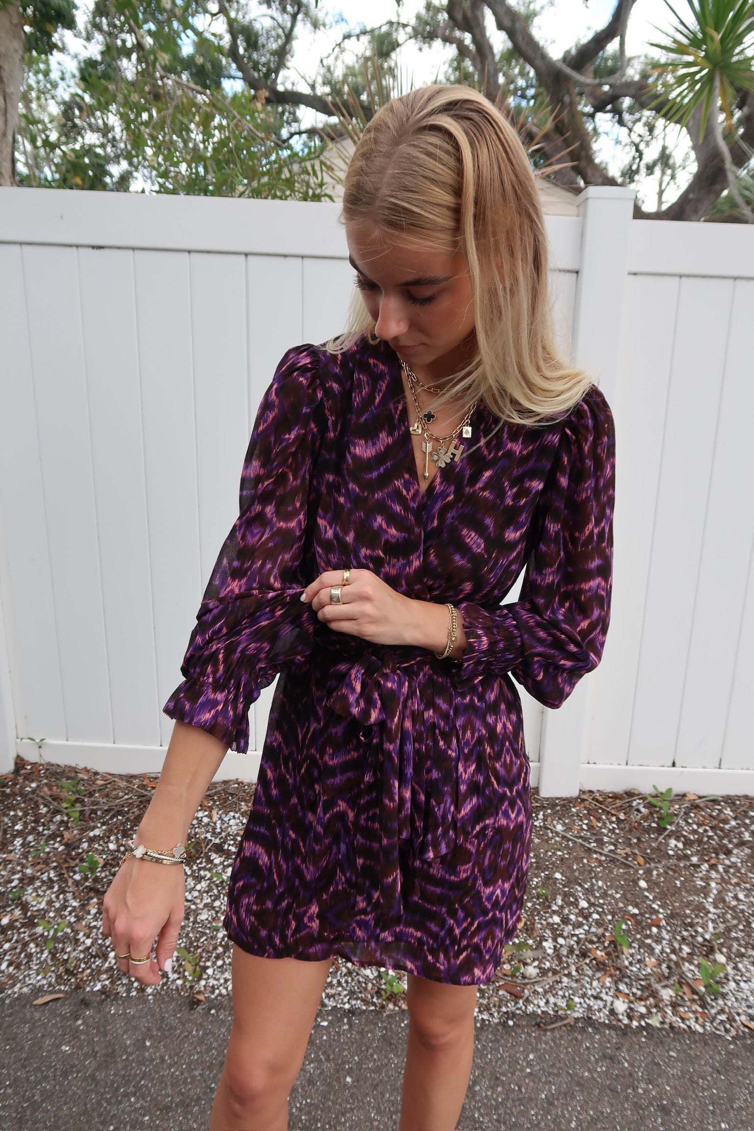 Woman wearing a purple patterned dress standing outdoors with a white fence and trees in the background. sleeve detail