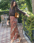 A woman posing outdoors on a brick patio path next to a green hedge, wearing a dark brown cropped jacket, a rust-red and beige floral print maxi skirt, and tan heeled sandals, while carrying a brown leather shoulder bag.