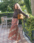 A woman stands outdoors on a brick patio wearing a dark brown cropped jacket, a long rust-red and beige floral print skirt, and carrying a tan shoulder bag.