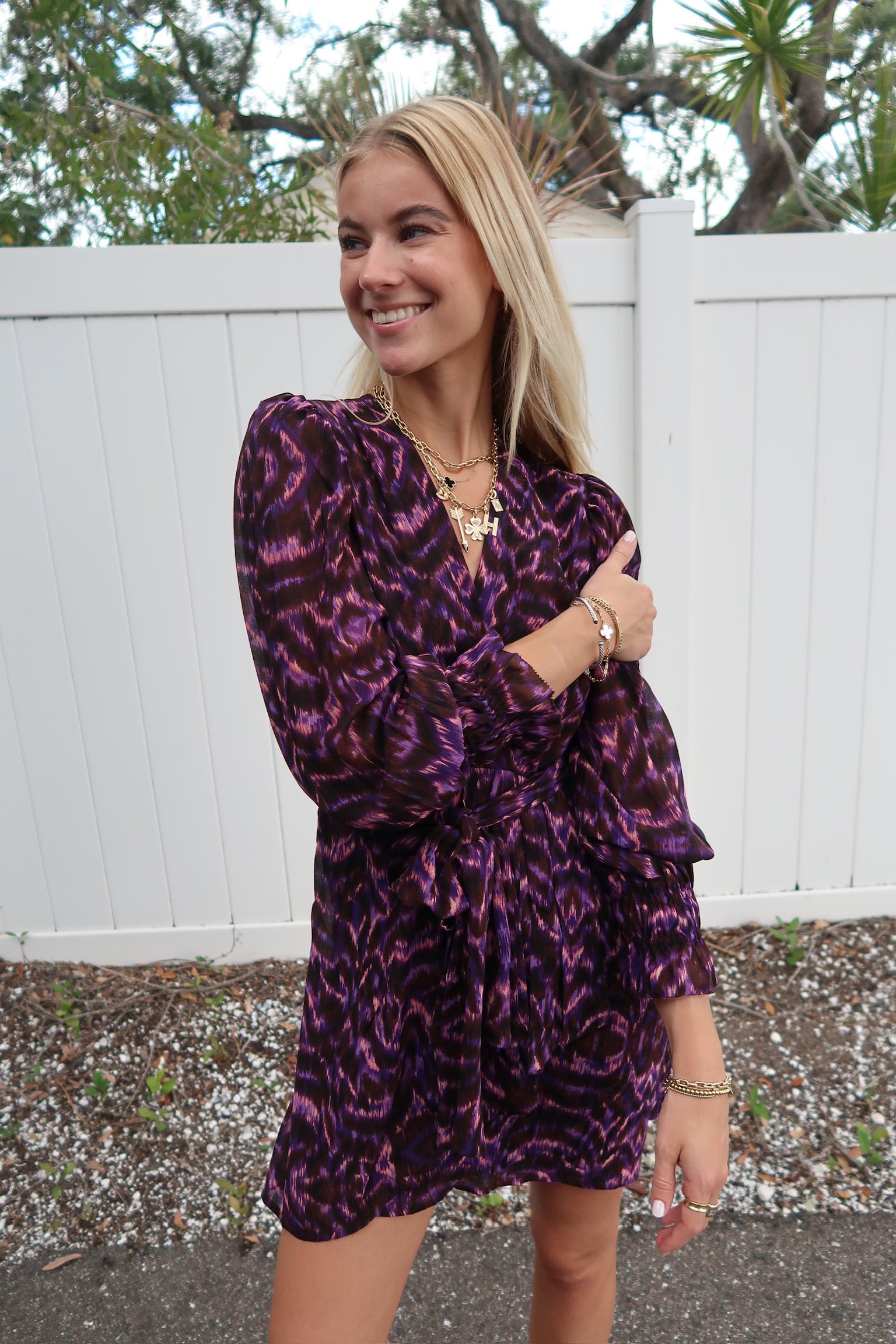 Woman wearing a purple patterned dress outdoors with a white fence and trees in the background.