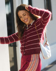 A woman wearing a fuzzy burgundy and pink striped zip-up sweater and matching striped pants, carrying a white shoulder bag, posing outdoors.