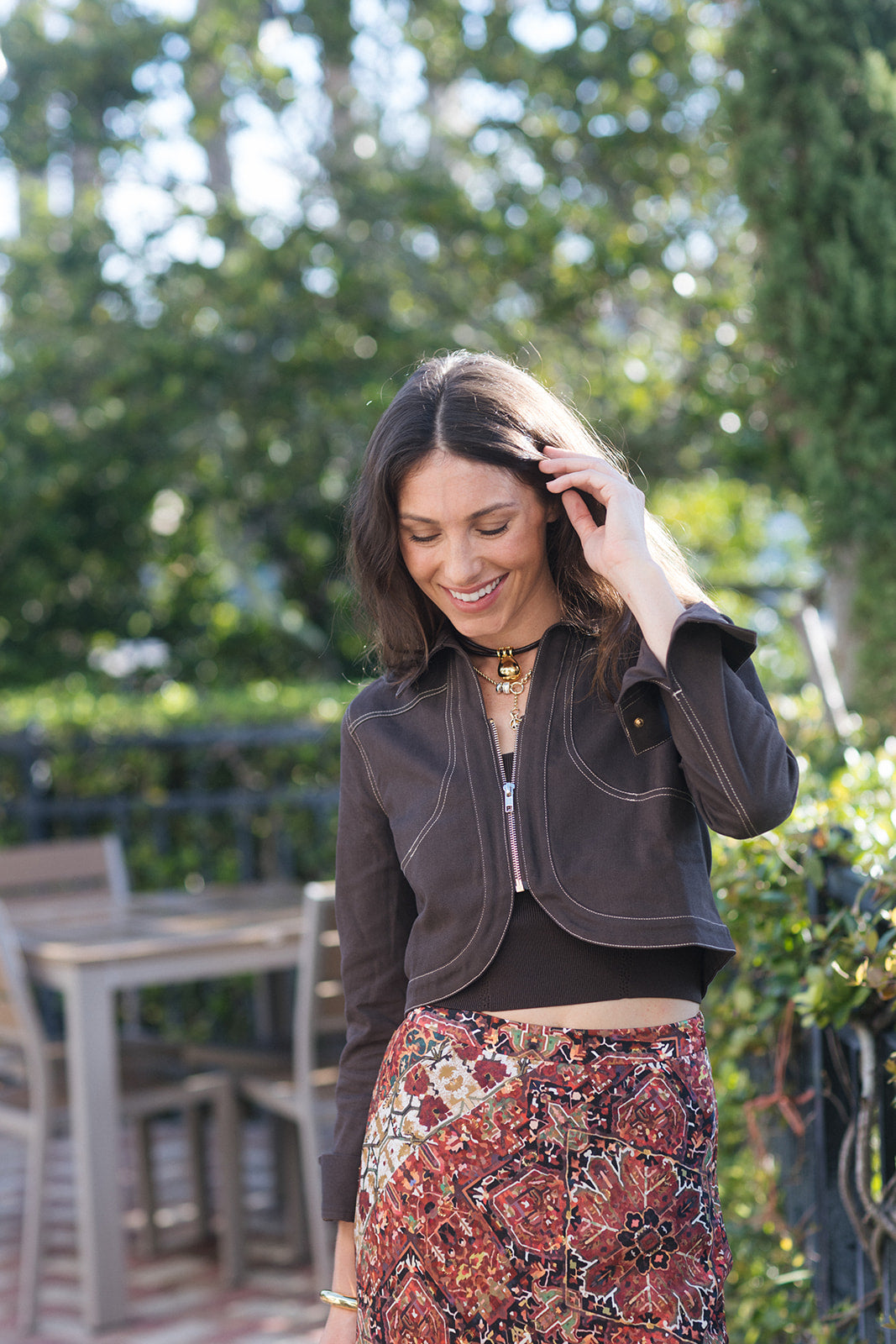 A woman wearing a dark brown cropped zip-up jacket and a red, brown, and black patterned skirt, standing outdoors in front of a blurred green garden background.
