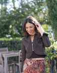 A woman wearing a dark brown cropped zip-up jacket and a red, brown, and black patterned skirt, standing outdoors in front of a blurred green garden background.