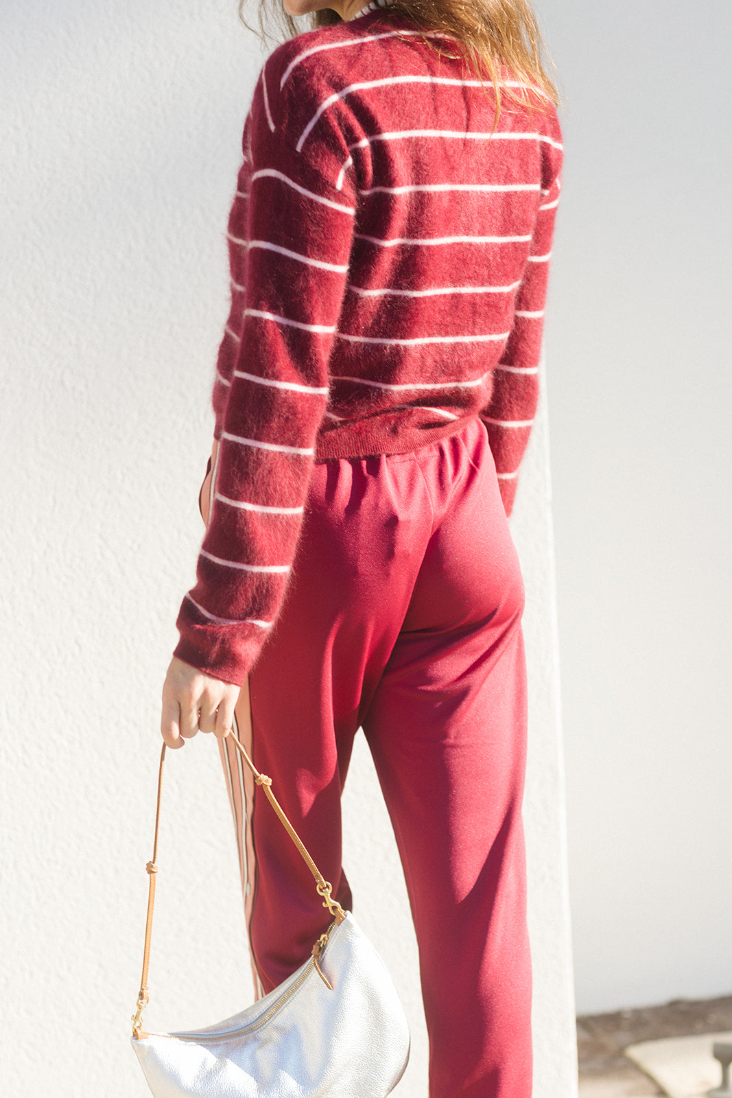 A woman wearing a fuzzy red and white striped long-sleeve sweater and matching red pants, holding a white handbag with a gold strap, standing against a white wall.
