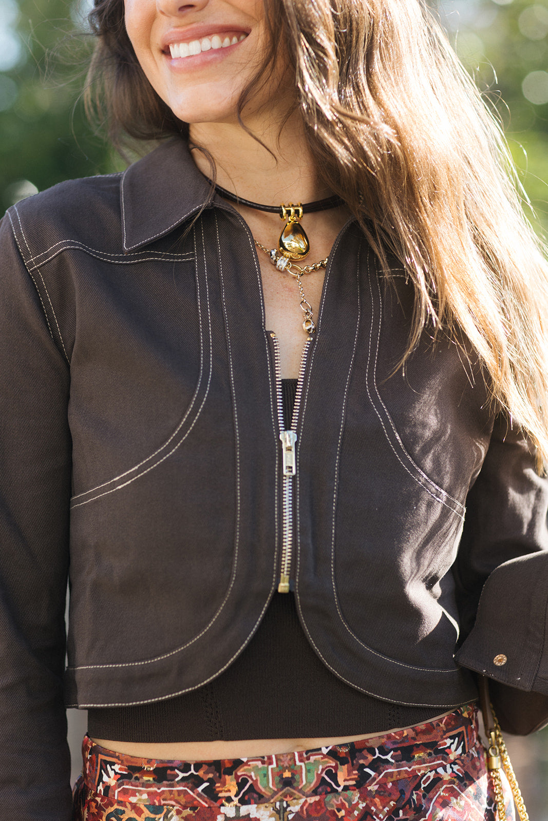A woman posing outdoors on a brick patio path next to a green hedge, wearing a dark brown cropped jacket, a rust-red and beige floral print maxi skirt, and tan heeled sandals, while carrying a brown leather shoulder bag, close up 