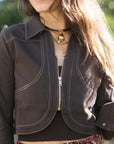 A woman posing outdoors on a brick patio path next to a green hedge, wearing a dark brown cropped jacket, a rust-red and beige floral print maxi skirt, and tan heeled sandals, while carrying a brown leather shoulder bag, close up 