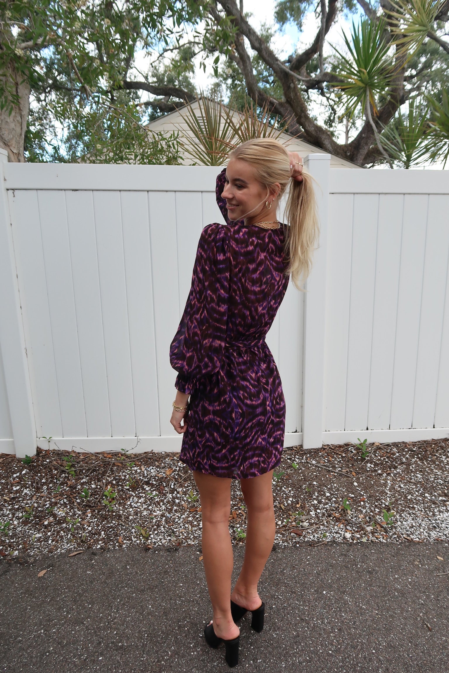 Woman wearing a purple dress with a pattern, standing in front of a white fence. full body back