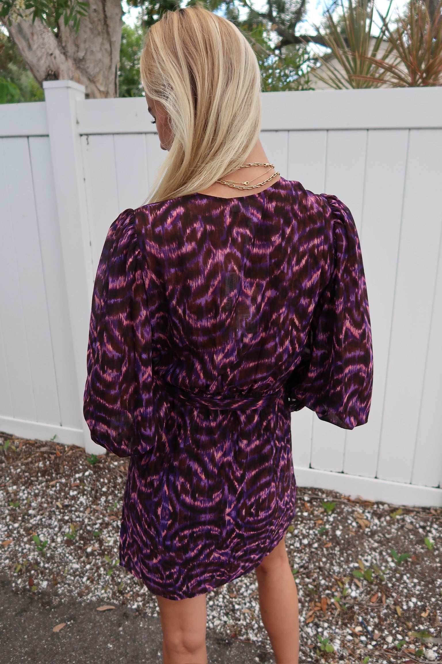 Person wearing a purple and black patterned dress with a white fence and trees in the background. close up back