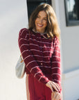 A woman smiling outdoors wearing a burgundy cardigan sweater with thin white horizontal stripes and matching burgundy trousers, carrying a white shoulder bag.