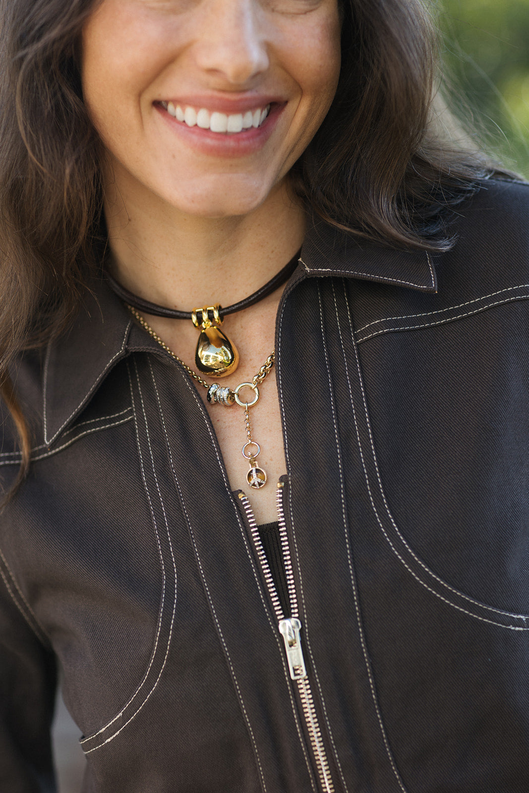 A close-up lifestyle shot of a woman wearing layered necklaces, including a black choker with a gold teardrop pendant and a gold beaded chain, paired with a dark brown jacket.
