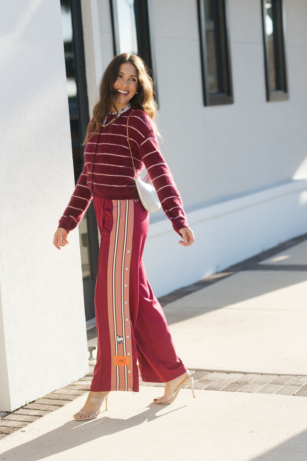 A woman walking outdoors wearing a burgundy and maroon striped long-sleeve top with matching wide-leg pants featuring vertical stripes, paired with nude high heels and a white shoulder bag.