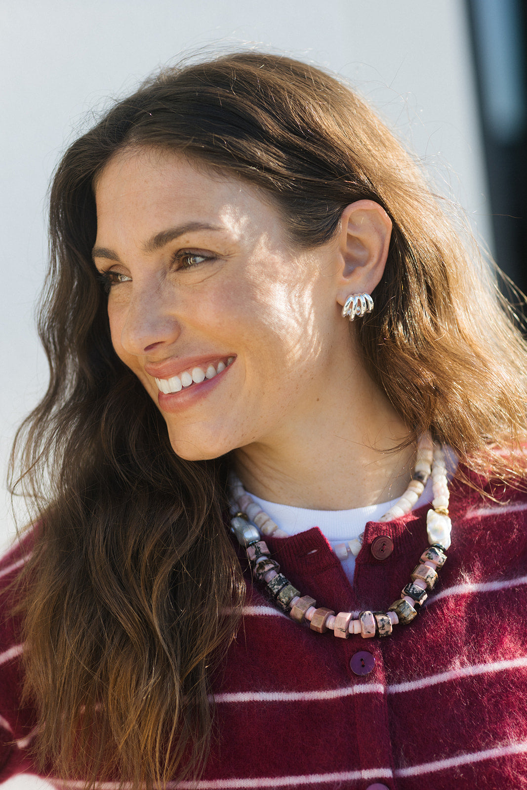 A smiling woman with long brown hair wearing a maroon and white striped knit top, a chunky multi-colored beaded necklace, and a silver-toned beaded hoop earring, photographed outdoors in bright sunlight.