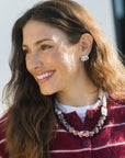 A smiling woman with long brown hair wearing a maroon and white striped knit top, a chunky multi-colored beaded necklace, and a silver-toned beaded hoop earring, photographed outdoors in bright sunlight.