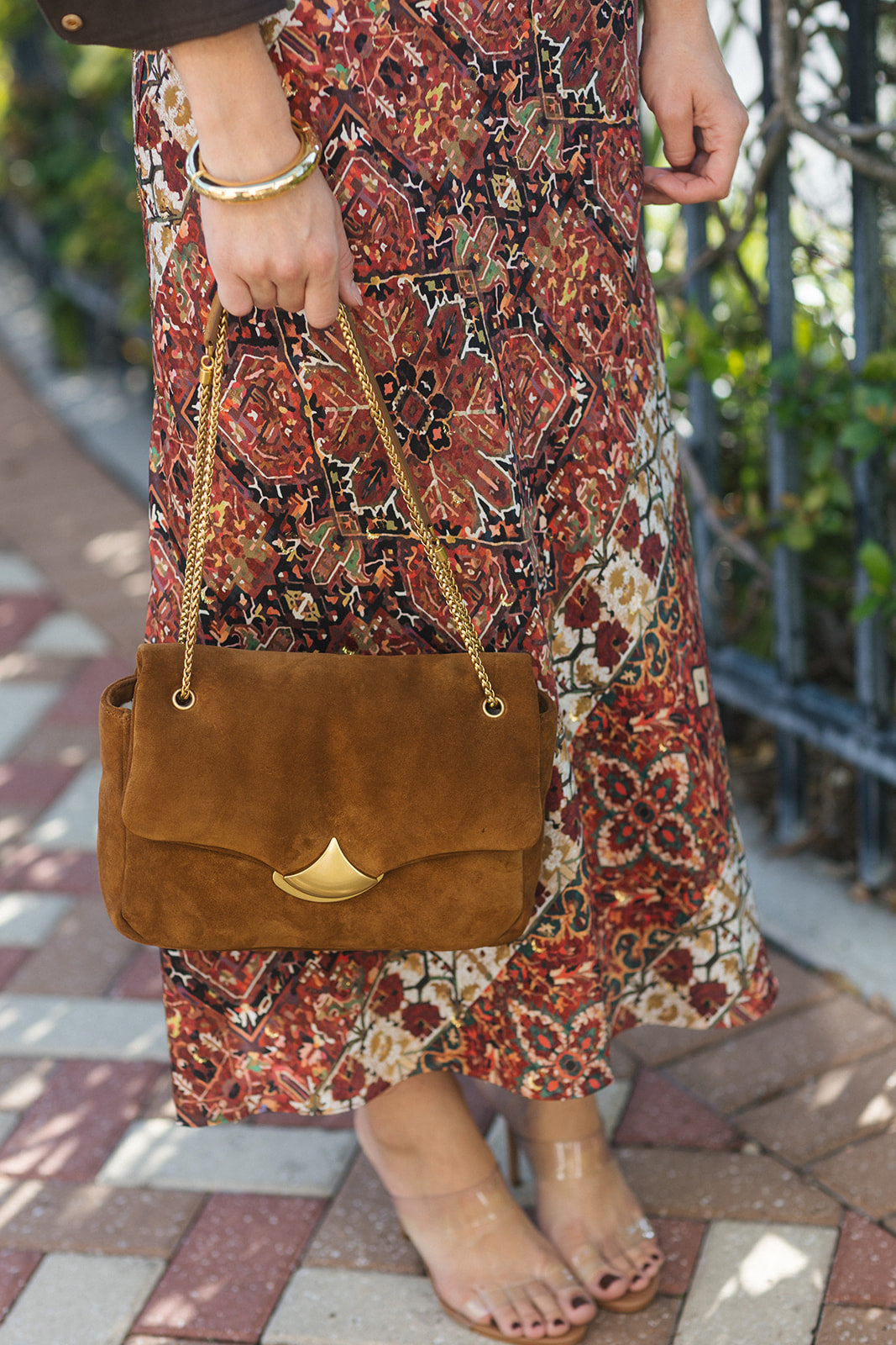 A person outdoors holding a brown suede shoulder bag with a gold chain strap and a gold triangular front accent. The person is wearing a long rust-red patterned maxi skirt and clear high-heeled sandals.