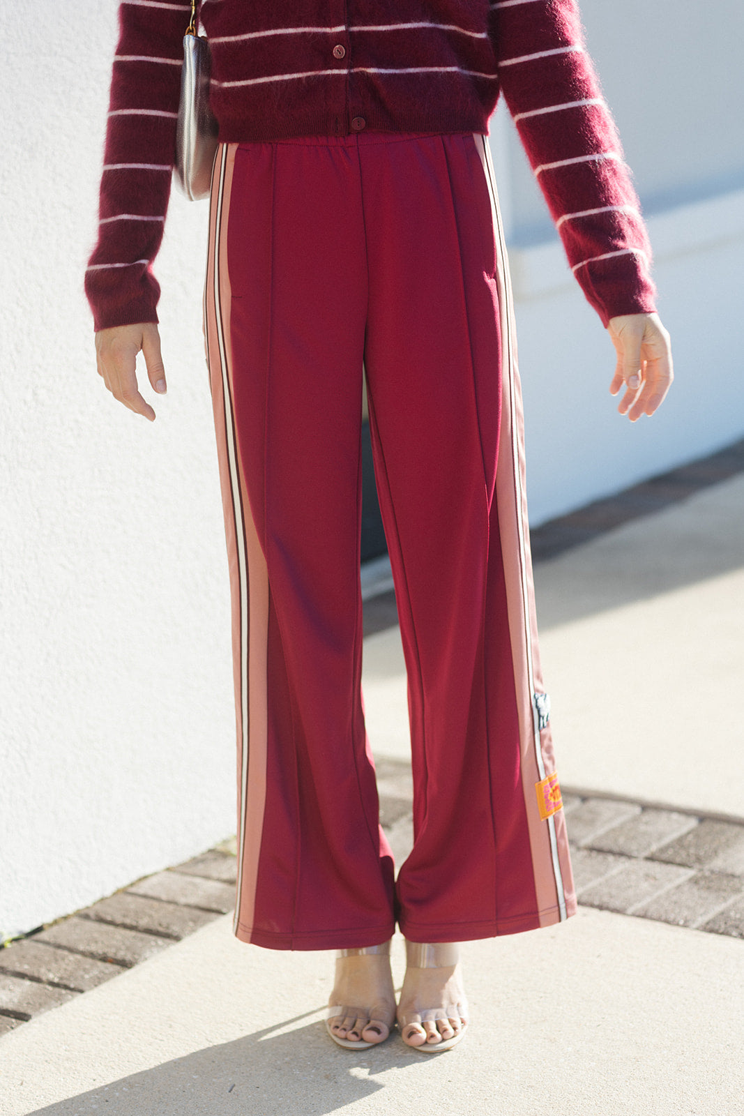 A person wearing burgundy wide-leg pants with beige side stripes, paired with a matching burgundy and white striped cardigan, standing outdoors on a paved surface.