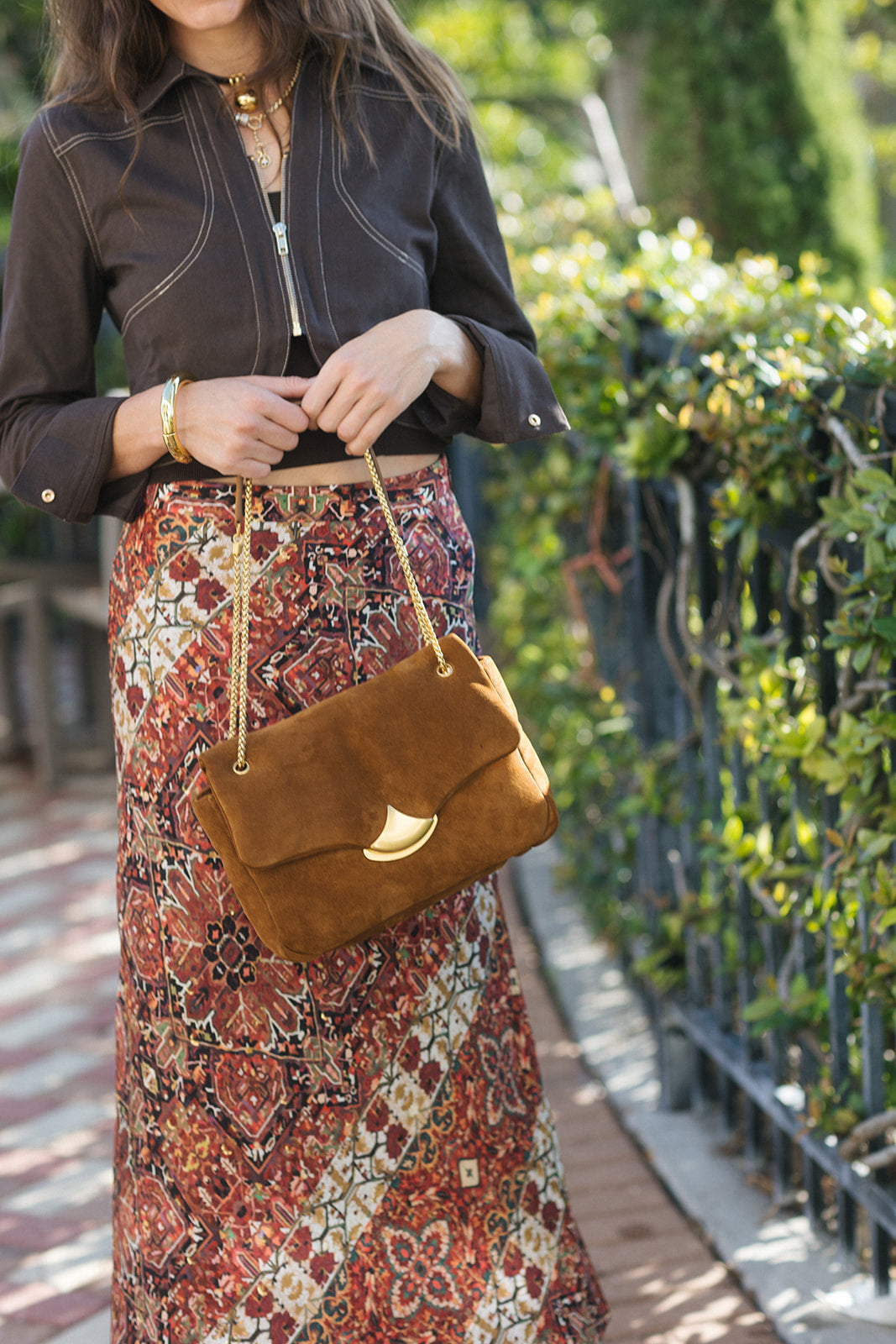 A woman outdoors wearing a dark brown cropped jacket and a long rust-red patterned skirt, carrying a brown suede flap shoulder bag with a gold-tone crescent moon detail and chain strap.