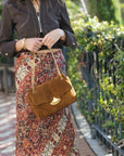 A woman outdoors wearing a dark brown cropped jacket and a long rust-red patterned skirt, carrying a brown suede flap shoulder bag with a gold-tone crescent moon detail and chain strap.