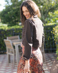 A woman wearing a dark brown cropped denim jacket and a rust-orange patterned midi skirt, standing in an outdoor garden patio setting.