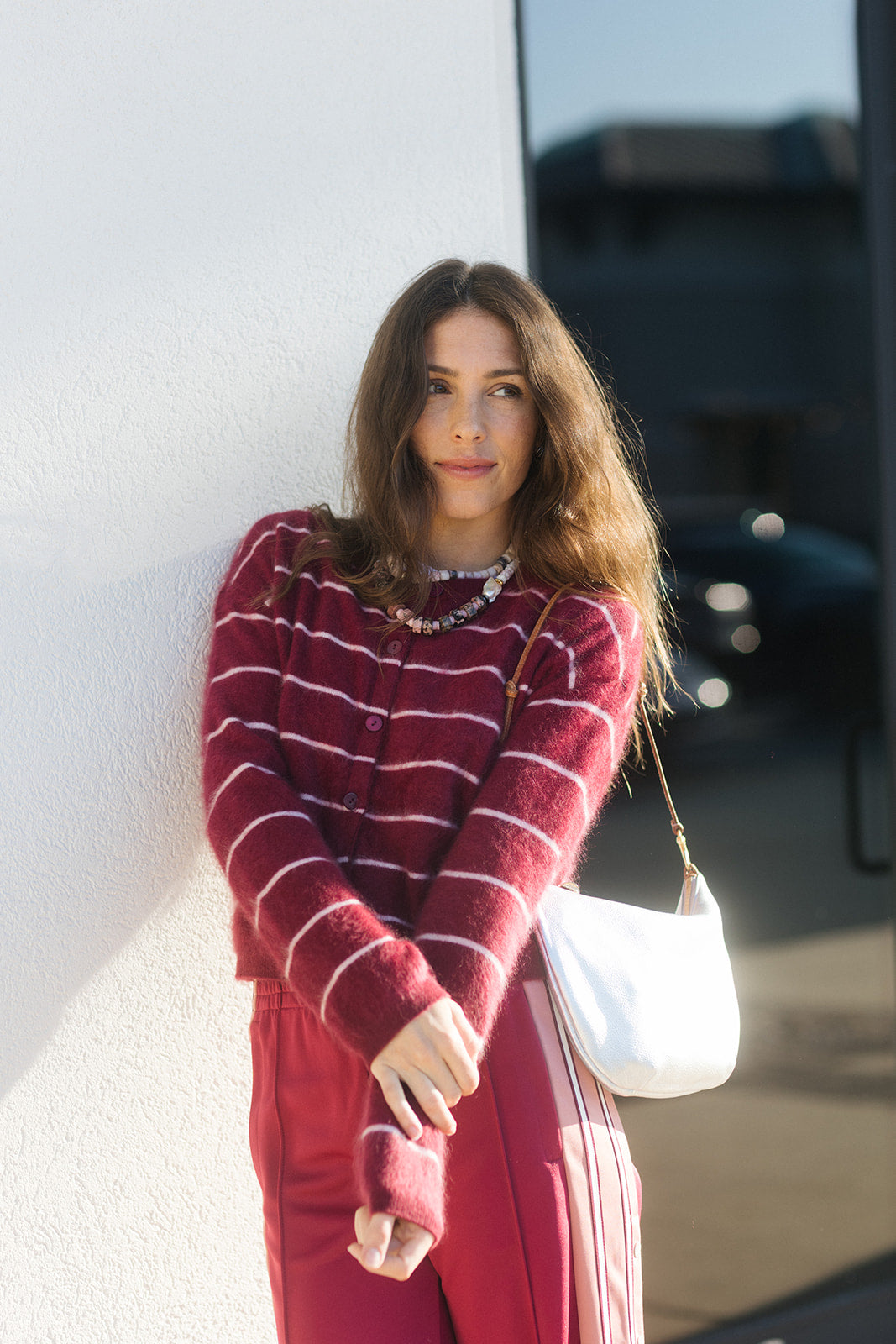 A woman wearing a burgundy and white striped knit cardigan and matching burgundy pants, leaning against a white wall outdoors. She is carrying a white shoulder bag.