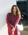 A woman wearing a burgundy and white striped knit cardigan and matching burgundy pants, leaning against a white wall outdoors. She is carrying a white shoulder bag.
