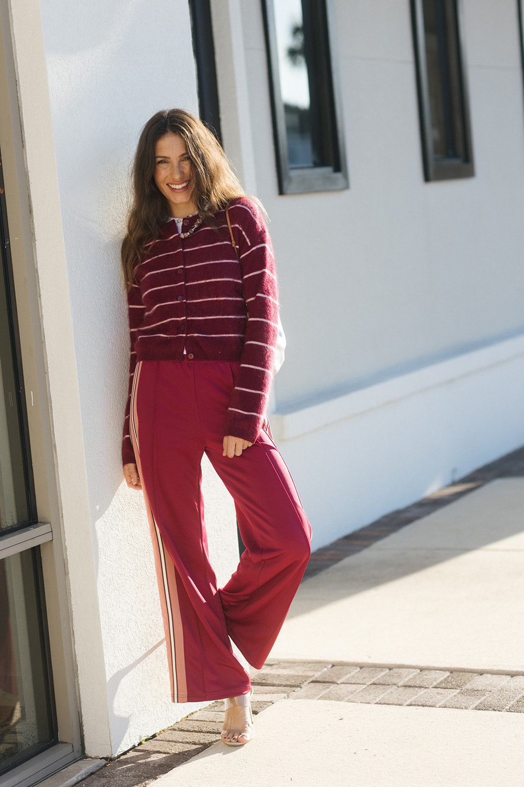A woman stands outdoors against a white building wall wearing a burgundy and white striped cropped zip-up cardigan with matching wide-leg burgundy pants featuring thin white side stripes.