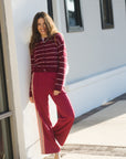 A woman stands outdoors against a white building wall wearing a burgundy and white striped cropped zip-up cardigan with matching wide-leg burgundy pants featuring thin white side stripes.