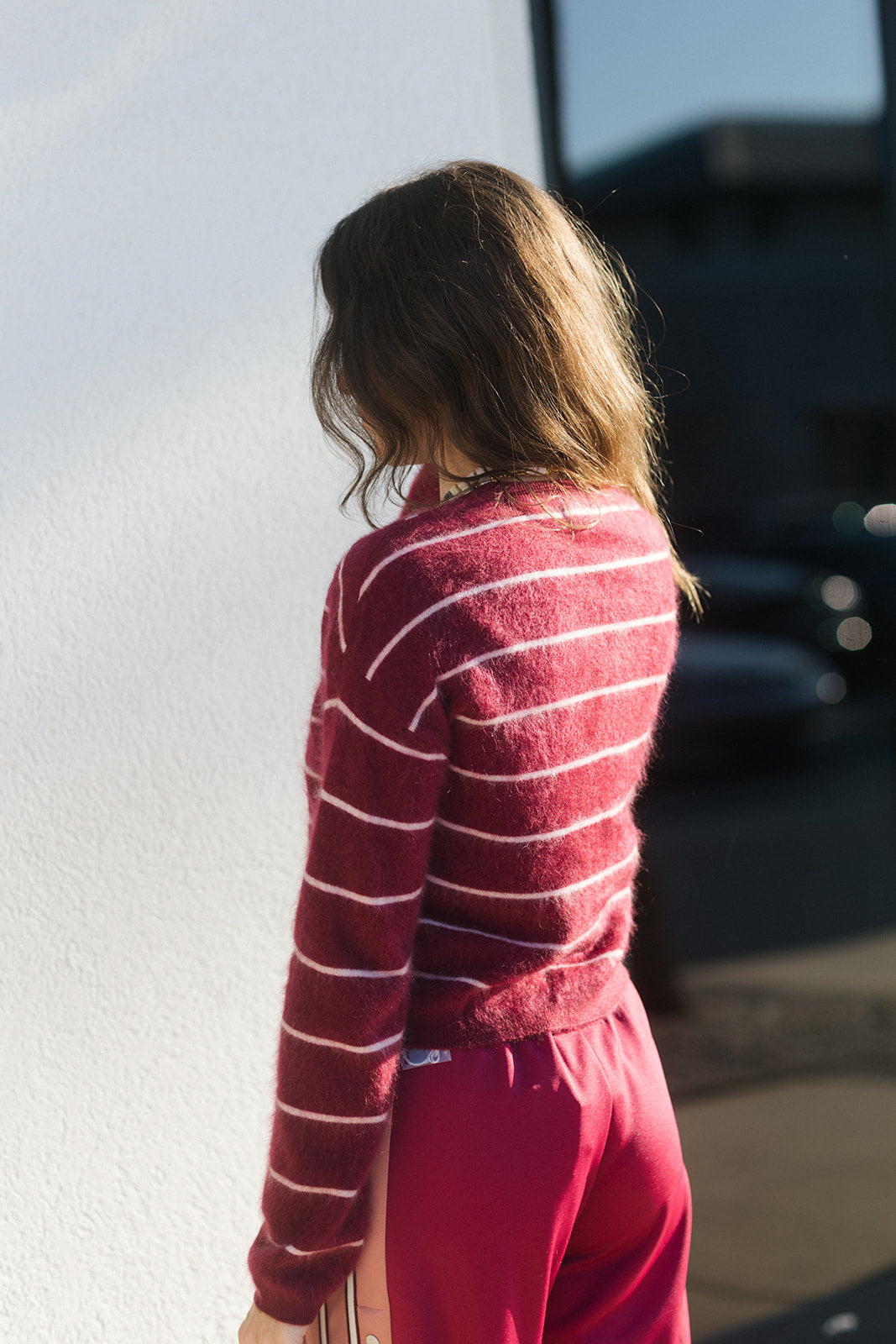 A woman shown from behind wearing a fuzzy long-sleeve knit sweater with thin white horizontal stripes on a deep red base, paired with bright pink pants, standing outdoors against a white wall.