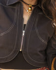Close-up of a person wearing a dark brown cropped jacket with a half-zip front, contrast white stitching, and layered gold necklaces.