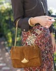 A woman wearing a dark brown jacket and a red and beige patterned skirt carries a brown suede shoulder bag with a gold-tone crescent clasp and chain strap.