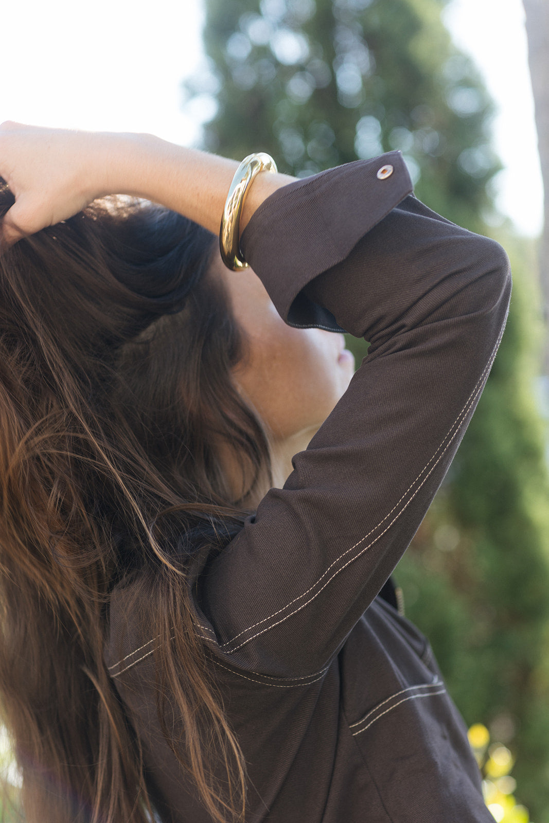 A woman with long brown hair wearing a dark brown long-sleeve shirt with white contrast stitching and a gold bangle, photographed outdoors with a blurred green background.