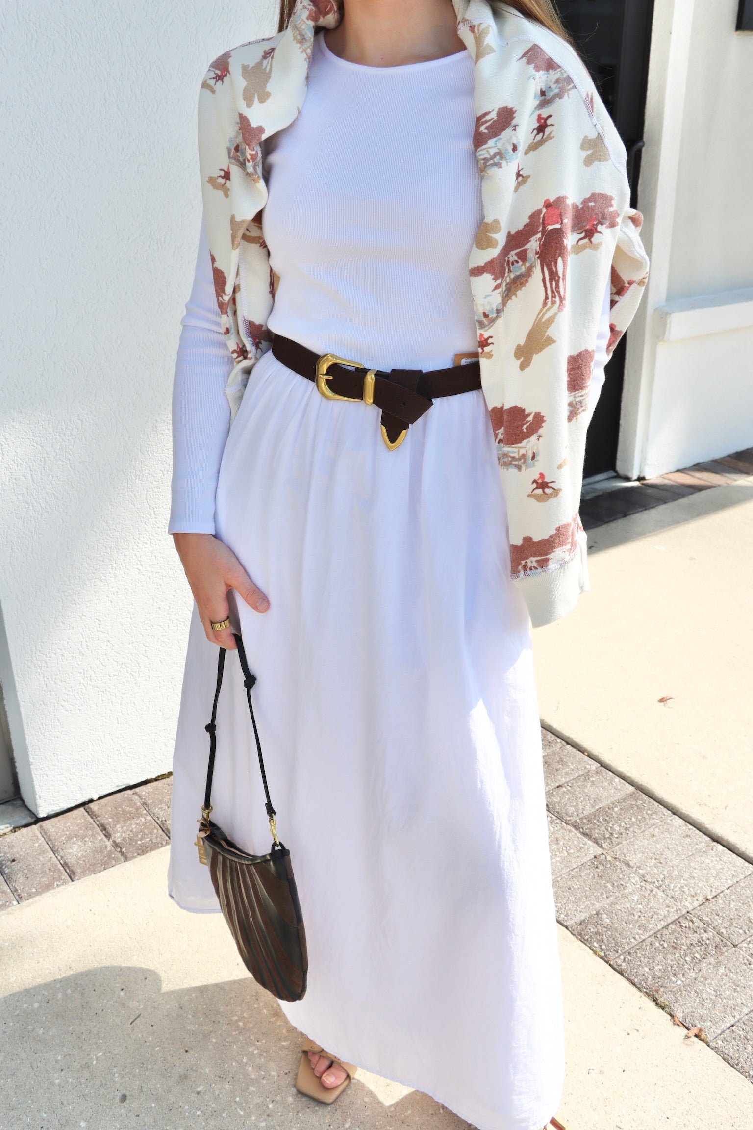 Person wearing a white dress with a floral cardigan and black belt, holding a brown bag.