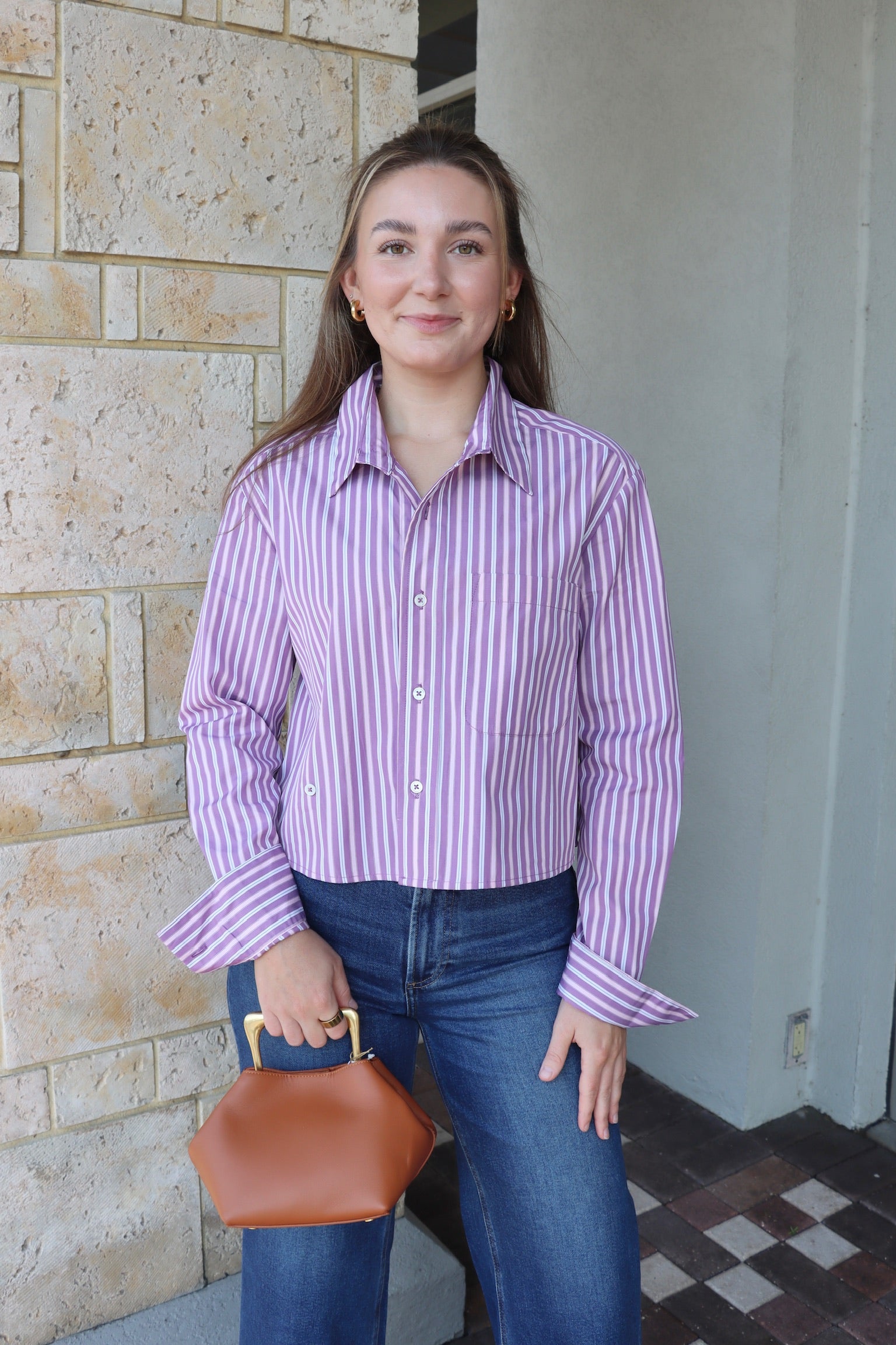 Woman wearing a purple striped shirt and blue jeans, holding a brown handbag, standing against a stone wall.