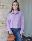 Woman wearing a purple striped shirt and blue jeans, holding a brown handbag, standing against a stone wall.
