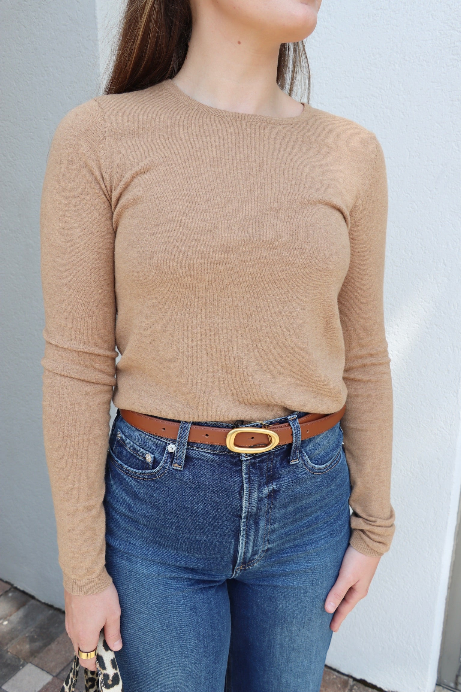 Person wearing a beige long-sleeve top and blue jeans with a belt, standing against a light-colored wall.