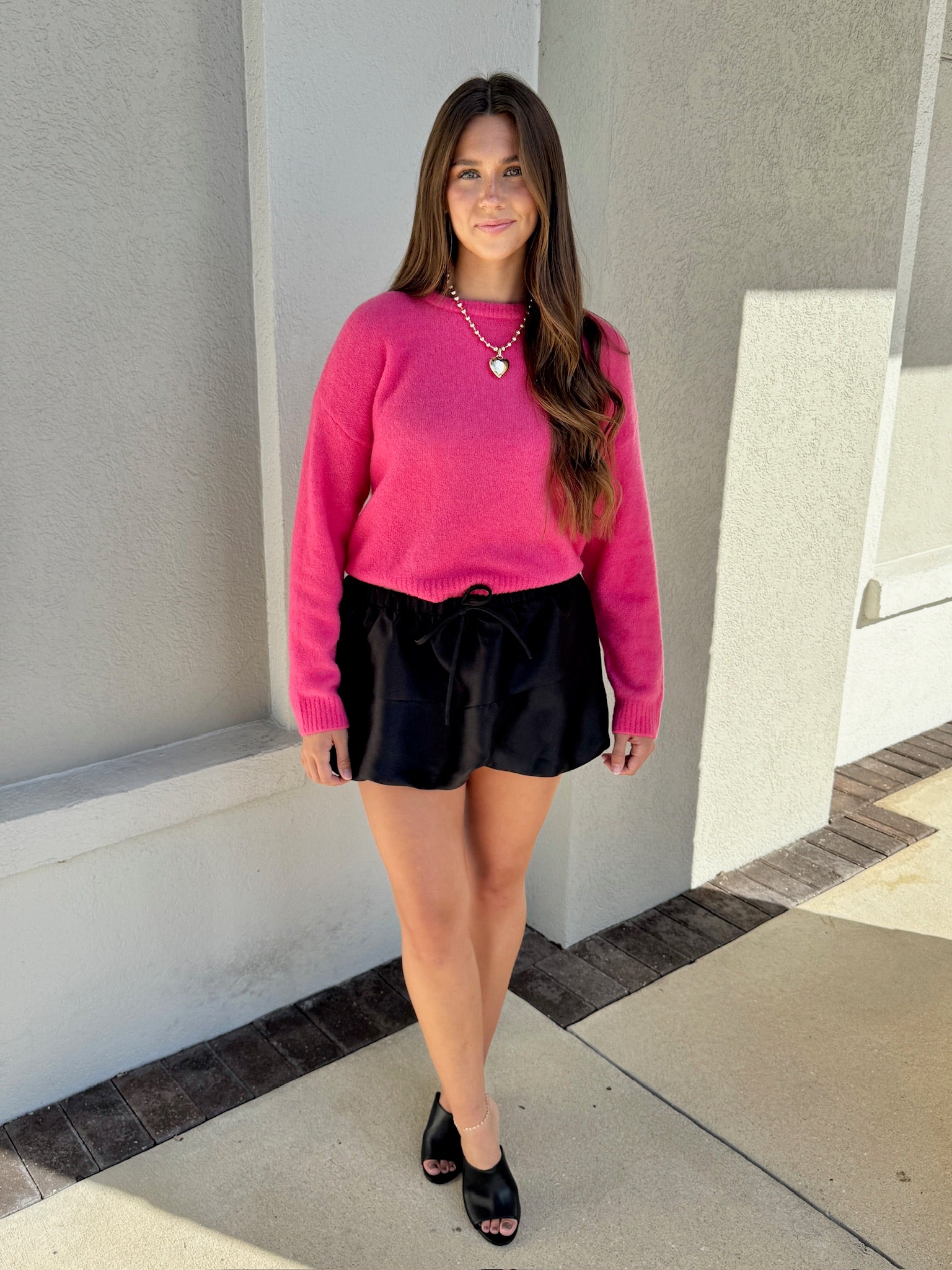 Woman wearing a pink sweater and black shorts standing against a light-colored wall.