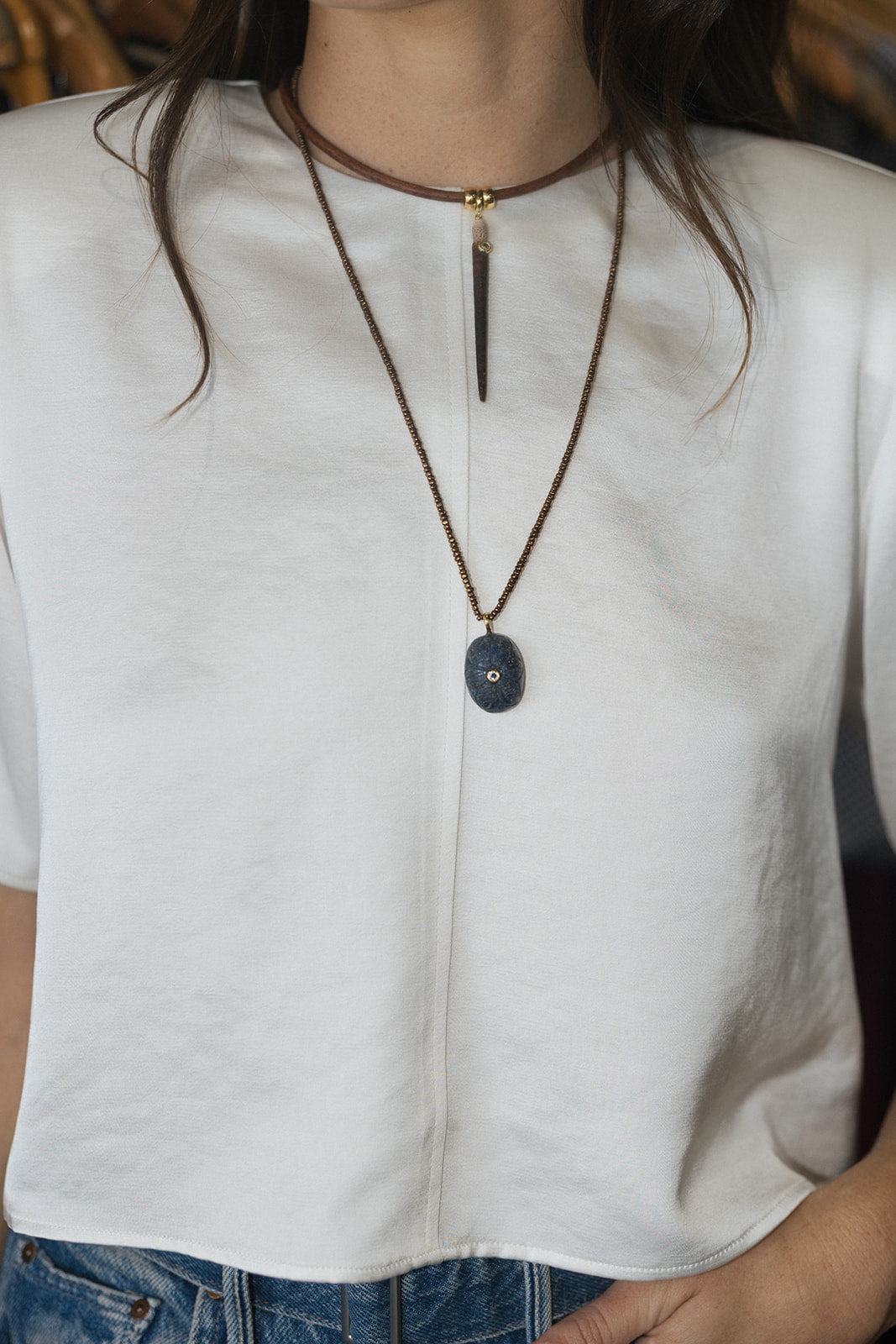 A close-up of a person wearing a white short-sleeve top and blue jeans, styled with a layered necklace featuring a dark gray oval pendant and a brown cord choker.