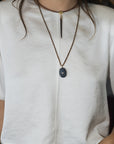 A close-up of a person wearing a white short-sleeve top and blue jeans, styled with a layered necklace featuring a dark gray oval pendant and a brown cord choker.