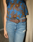 A person wearing a sleeveless blue top with a brown and gold floral pattern, paired with light-wash blue jeans and a brown belt with a gold buckle.