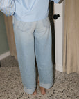 A person shown from behind wearing light-wash wide-leg denim jeans with rolled cuffs and a light blue top, standing barefoot on a speckled floor in an indoor room.
