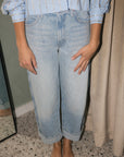 A person wearing light-wash cropped wide-leg denim jeans with rolled cuffs, paired with a light blue embellished blouse, standing barefoot indoors.