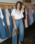 A woman posing in a clothing boutique wearing a white short-sleeve crop top, high-waisted wide-leg blue jeans, and holding a small brown textured handbag.