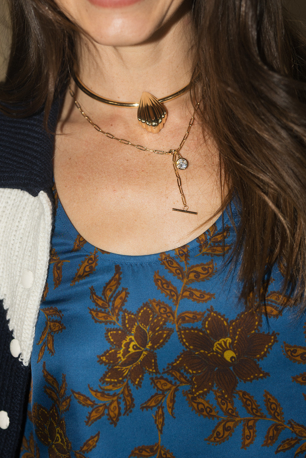 A close-up of a model wearing layered gold necklaces, including a choker with a shell pendant and a delicate chain with a T-bar and small round charm, over a blue floral top.